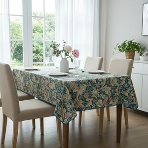 May include: A rectangular tablecloth with a floral pattern in teal, beige, and pink covers a wooden dining table. The table is set with plates, glasses, and a vase of flowers. Beige chairs surround the table, creating a dining room setting.