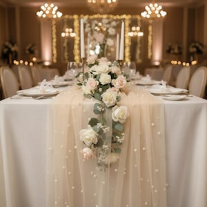 May include: An elegant table setting, ideal for a wedding or special occasion. The white tablecloth is adorned with a pearl-studded runner. A floral centrepiece of white and pink roses, candles, and place settings creates a sophisticated atmosphere for the event.