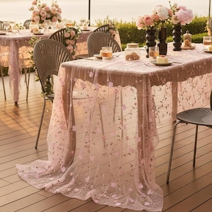 Può includere: Una tovaglia trasparente rosa chiaro con delicati motivi floreali ricamati in tonalità rosa e verde. Il tessuto ricade sui bordi di un tavolo rettangolare, allestito con vasi di vetro, composizioni floreali e piccoli piatti.
