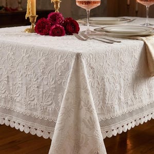 Pode incluir: Uma toalha de mesa branca bordada com um desenho floral. A toalha de mesa tem uma borda de renda com vieiras. Flores vermelhas, velas e vidro estão sobre a mesa, sugerindo um ambiente de jantar formal.