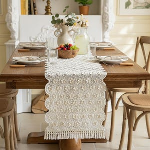 Puede incluir: Una mesa de comedor de madera preparada para una comida, con un camino de mesa de encaje bordado de color crema, con borde festoneado y flecos. La mesa está puesta con platos, vasos y cubiertos con mango de madera. También hay un cuenco de madera con fruta y un jarrón con flores.
