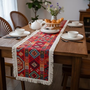 Op de afbeelding: Een rode tafelloper met een geometrisch patroon en witte franjes. De loper ligt op een houten tafel die gedekt is voor een maaltijd, met witte borden, kommen en bestek. Een broodmand en een theepot zijn ook aanwezig.