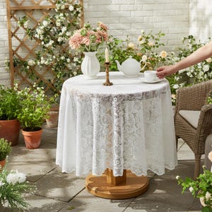 Puede incluir: Una mesa redonda al aire libre con un mantel de encaje blanco, una tetera y un jarr&oacute;n con flores. Una mano alcanza una taza de t&eacute;. La mesa est&aacute; sobre una base de madera, rodeada de plantas y una pared de ladrillos.