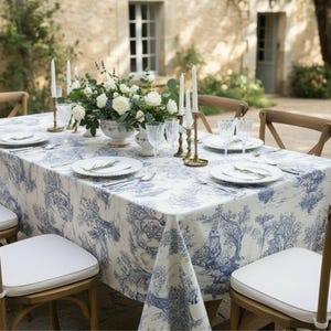 Op de afbeelding: Een buitentafel gedekt met een blauw-wit toile de Jouy tafelkleed. Elke plaatsing omvat witte borden, bestek en heldere glazen. Een bloemstuk en kaarsen completeren de elegante sfeer. Houten stoelen omringen de tafel.