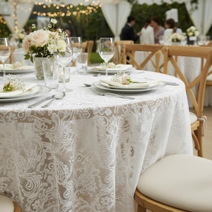 Puede incluir: Una mesa redonda preparada para una recepci&oacute;n de boda, cubierta con un mantel de encaje blanco. La mesa est&aacute; puesta con platos, cubiertos y copas de vino. Un centro de mesa floral y peque&ntilde;os acentos florales se suman a la elegante decoraci&oacute;n.