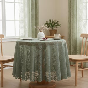 Puede incluir: Un mantel redondo de encaje verde salvia cubre una mesa de madera, con un jarr&oacute;n blanco, un plato de cerezas y una taza de t&eacute;. Dos sillas de madera flanquean la mesa, con cortinas verdes transparentes en el fondo.