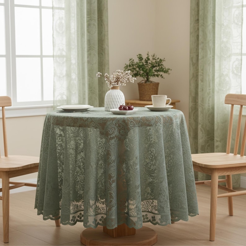 Puede incluir: Un mantel redondo de encaje verde salvia cubre una mesa de madera, con un jarr&oacute;n blanco, un plato de cerezas y una taza de t&eacute;. Dos sillas de madera flanquean la mesa, con cortinas verdes transparentes en el fondo.