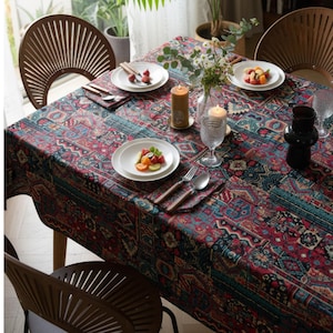Puede incluir: Una mesa de comedor con un mantel estampado en tonos rojos, azules y turquesas. Platos blancos con ensalada de frutas, cubiertos y servilletas están dispuestos. Velas y un jarrón de flores decoran la mesa. Sillas de madera rodean la mesa.