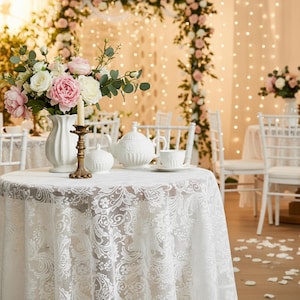 Puede incluir: Una mesa redonda preparada para una ocasi&oacute;n especial, cubierta con un mantel de encaje blanco. Un juego de t&eacute; de porcelana blanca, un jarr&oacute;n de flores y un candelabro est&aacute;n sobre la mesa. El fondo presenta arcos florales y luces de cadena.