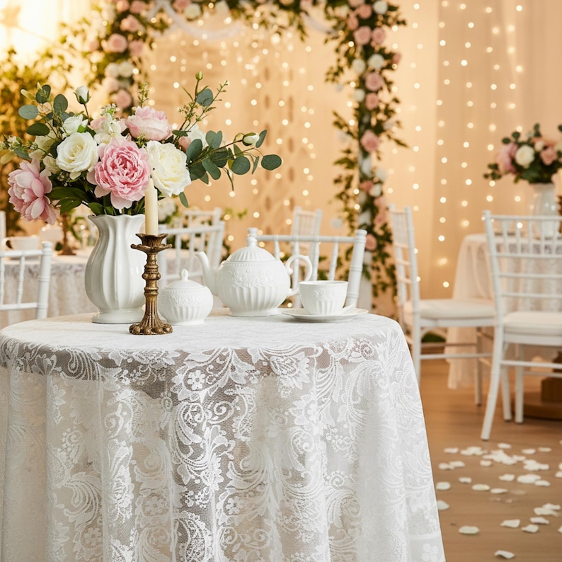 Puede incluir: Una mesa redonda preparada para una ocasi&oacute;n especial, cubierta con un mantel de encaje blanco. Un juego de t&eacute; de porcelana blanca, un jarr&oacute;n de flores y un candelabro est&aacute;n sobre la mesa. El fondo presenta arcos florales y luces de cadena.