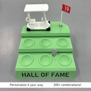 Op de afbeelding: Een groene golf-thema displaystandaard met een witte golfkar, een rode vlag met het nummer 19 en zes ronde houders. De basis leest "HALL OF FAME". De tekst "Personalize it your way" en "200+ combinations!" zijn ook aanwezig.