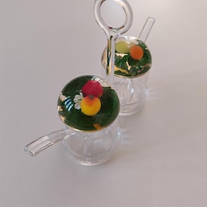 Peut inclure: Deux tasses à thé en verre transparent avec une poignée et un petit bec verseur. Chaque tasse a un dessus rond en verre vert avec des fruits et des feuilles incrustés. La poignée est une boucle claire et circulaire. Les tasses sont sur une surface blanche.