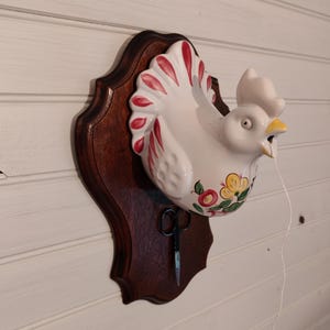 May include: A white ceramic rooster wall decor with red and yellow floral accents, mounted on a dark brown wooden plaque. A pair of small scissors hangs below the rooster. A white string extends from the rooster's beak.