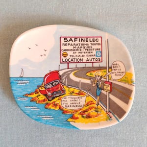 Peut inclure: Assiette décorative en céramique avec une scène de dessin animé. Une voiture rouge est accidentée sur une route côtière, avec une femme en robe bleue. Le texte comprend "SAFINELEC" et "LOCATION AUTOS".