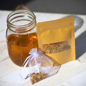 Può includere: Un barattolo di vetro trasparente pieno di liquido ambrato, un sacchetto di carta marrone con una finestra trasparente e un piccolo sacchetto di rete bianco. Il sacchetto e la busta contengono erbe e spezie essiccate.