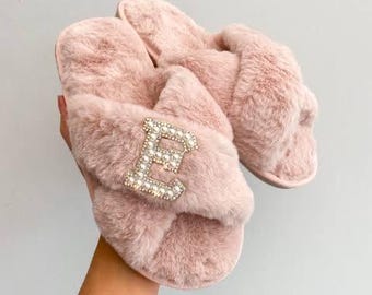 May include: A pair of pink, fluffy slippers with a large, embellished letter "E" on the top. The slippers have a soft, plush texture and a cross-strap design. The letter "E" is encrusted with small, shiny stones.