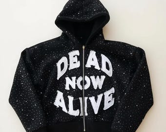 May include: Black zip-up hoodie with a starry pattern and the words "DEAD NOW ALIVE" in large, white, textured lettering. The hoodie has a hood and a silver zipper.