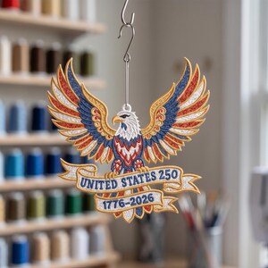 Może przedstawiać: Haftowana patriotyczna ozdoba w kształcie orła z rozpostartymi skrzydłami, w kolorach czerwonym, białym i niebieskim. Na bannerze napis "UNITED STATES 250 1776-2026". Ozdoba zawieszona na srebrnym haczyku.