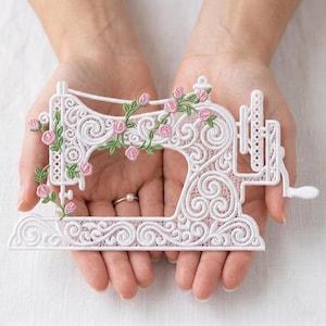 May include: A white, decorative sewing machine featuring detailed scrollwork and pink rose embellishments. The machine is decorated with a floral garland and held in open hands. A delicate craft item, perfect for sewing enthusiasts.