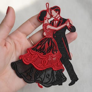 Puede incluir: Parche bordado que representa a una pareja bailando. La mujer lleva un vestido rojo y negro, mientras que el hombre viste un esmoquin negro. El parche está detallado con puntadas intrincadas, mostrando un tema de baile de salón.