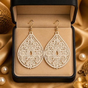 Pode incluir: Um par de brincos em forma de lágrima na cor creme, com um bordado intrincado semelhante a renda, apresentando padrões de espirais e motivos florais. Os brincos estão suspensos em ganchos de metal dourado dentro de uma caixa preta.