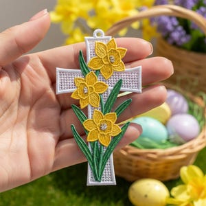 以下が含まれることがあります： 黄色いスイセンの花と緑の葉が刺繍された白い十字架のペンダント。十字架の上部には吊り下げ用の小さなループが付いています。花は詳細な黄色の花びらと質感のある中心部を持っています。