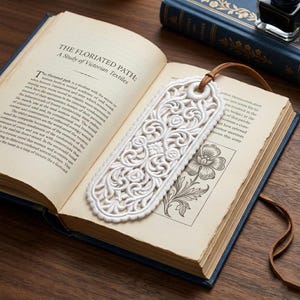 Puede incluir: Un marcapáginas blanco, con un diseño intrincado de flores, colocado sobre las páginas de un libro abierto. El marcapáginas tiene una correa de cuero adjunta. El libro tiene una cubierta azul y el título "THE FLORIATED PATH: A Study of Victorian Textiles".