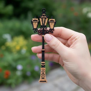 May include: An embroidered patch depicting a black and gold vintage street lamp. The lamp has three lanterns and ornate detailing. The patch is held in a hand against a blurred green backdrop.