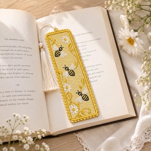 May include: A gold-colored bookmark with a yellow background, featuring embroidered bees and daisies. The bookmark is rectangular with a decorative border and a cream-colored tassel. It rests in an open book with text visible.