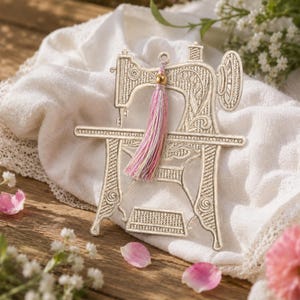 May include: A cream-coloured sewing machine ornament with detailed patterns. It has a pink, gold, and white tassel. The ornament rests on a white cloth with lace edging, with flowers and petals scattered around it.