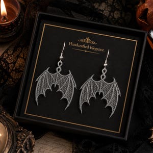 May include: A pair of silver-toned bat wing earrings displayed in a black box. The earrings are intricately designed with a detailed wing pattern. The box has the text "Handcrafted Elegance" in gold lettering.
