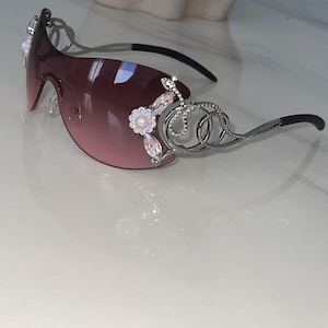 Puede incluir: Gafas de sol sin montura con una lente degradada rosa de una sola pieza. Las patillas de metal plateado están decoradas con un motivo floral con una perla central, cristales rosas de talla marquesa y un diseño de serpiente con pedrería.