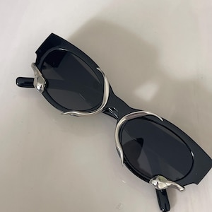 May include: Black sunglasses featuring dark oval lenses and a black frame. The front of the frame is accented with polished silver-tone metal hardware that curves around the outer edges of each lens, creating a distinct metallic decorative detail.