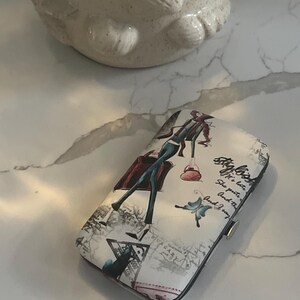 Peut inclure: Un étui à cartes à motifs blancs et noirs avec une illustration de mode d'une femme en tenue turquoise, un chapeau rouge et une valise rouge. L'étui a un fermoir argenté et le texte "Stylish".