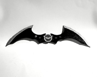 May include: A black bat-shaped throwing weapon with a metallic silver edge and a central circular emblem featuring a small bat silhouette. The object has a symmetrical design with three circular cutouts on each wing and a textured black finish.