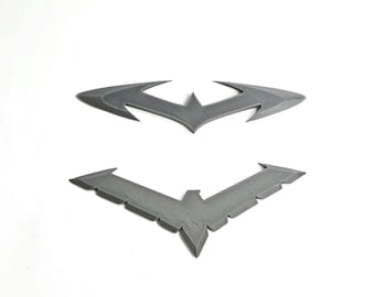 May include: Two gray, flat, decorative emblems. The top emblem is a stylized bat-shaped symbol with pointed wings. The bottom emblem is a V-shaped symbol with a central point and multiple serrated edges along the wings, resembling a bird motif.