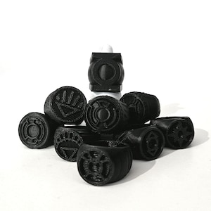 May include: A collection of matte black rings featuring various circular emblems and symbols on the face of each band. The rings are stacked together, displaying distinct geometric designs including star-like shapes and lantern-style icons.
