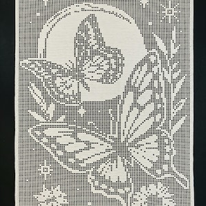 May include: A rectangular off-white crochet wall hanging featuring a grid-based pattern of two butterflies, a large circular moon, small stars, a floral sprig, and a flower. The intricate needlework creates a high-contrast design on the fabric.