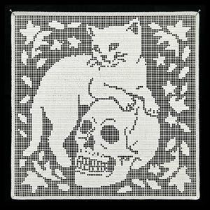 May include: A square white crochet doily featuring a filet pattern of a cat sitting atop a human skull, surrounded by a border of stylised leaves and small star shapes. The intricate needlework creates a high-contrast graphic design.