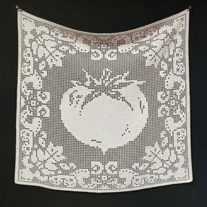 May include: A square, white, crocheted tablecloth featuring a large apple design at its centre. The apple is framed by a decorative border of floral and geometric patterns. The tablecloth is displayed against a black backdrop.
