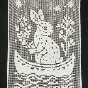 Könnte beinhalten: Weiß gehäkeltes Wandbehang mit einem Hasen in einem Boot, umgeben von Blumen- und Sternmotiven. Das Design ist mit einem gitterartigen Muster gestaltet, das einen strukturierten Effekt erzeugt. Der Hintergrund ist schwarz.