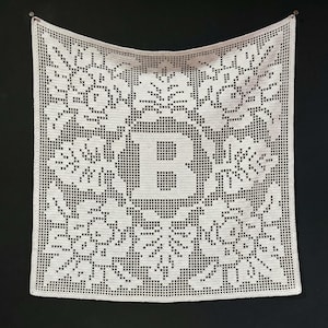 May include: A square, off-white crocheted blanket with a floral design and the letter "B" in the center. The blanket is made of a light-colored yarn and is displayed against a dark background.