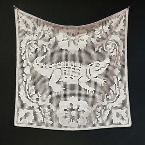May include: A square, off-white crocheted doily with a floral and bird design. The doily is suspended against a black background, showcasing the intricate openwork pattern. The design includes large flowers and stylized birds.
