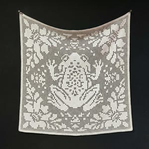 May include: A square, cream-coloured crocheted doily or wall hanging. The intricate design features floral and geometric patterns, with a central motif and a border of stylized flowers. It is suspended against a black background.
