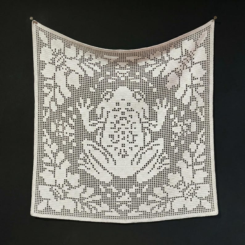 May include: A square, cream-coloured crocheted doily or wall hanging. The intricate design features floral and geometric patterns, with a central motif and a border of stylized flowers. It is suspended against a black background.