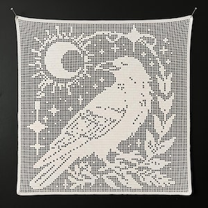 May include: A square white crochet wall hanging featuring a detailed filet pattern of a large bird in profile. The design includes a sun, a crescent moon, stars, and foliage elements surrounding the bird, all created with a grid-like stitch.