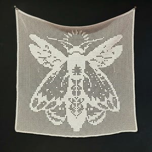 May include: A cream-colored, crocheted wall hanging featuring a large bee design. The bee is rendered in a detailed, openwork pattern against a square grid background. The piece is hung against a dark background.