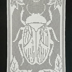 May include: White, rectangular, crocheted wall hanging with a detailed beetle design. The beetle is central, with wings and a head. The border features floral and leaf patterns. It is hung with a silver-coloured rod.