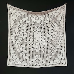 May include: A square, off-white crocheted piece featuring a central bee design surrounded by floral patterns. The openwork design is displayed against a dark background, showcasing the intricate details of the handmade textile.
