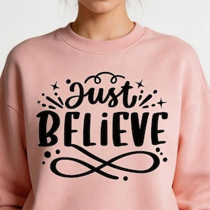 Puede incluir: Sudadera rosa claro con la frase "Just Believe" en letras negras. El diseño incluye estrellas y remolinos decorativos. La sudadera tiene cuello redondo y mangas largas.